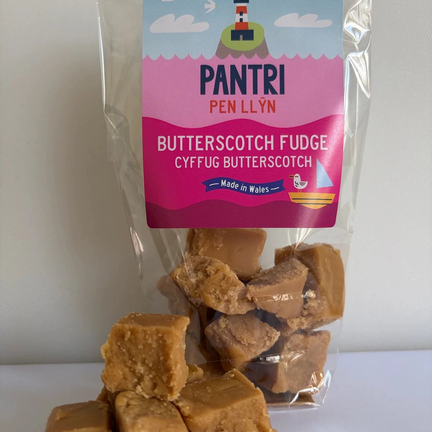 A clear plastic bag of Pantri Pen Llyn Butterscotch Fudge with several pieces of golden-brown fudge spilled in front on a white surface against a light gray background.