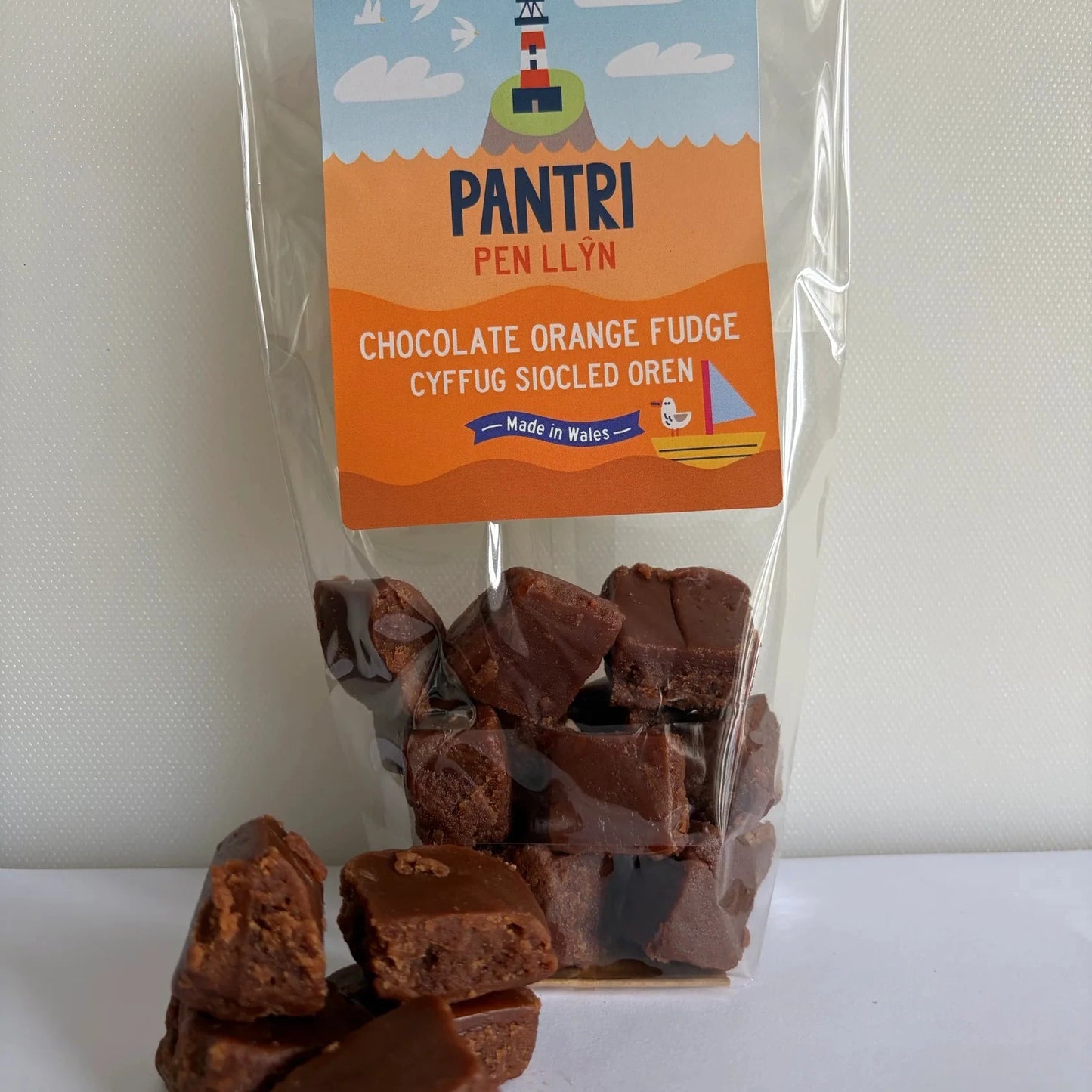 A clear stand-up pouch of Pantri Pen Llyn Chocolate Orange Fudge, with several pieces of fudge spilled in front on a white surface against a light gray wall.