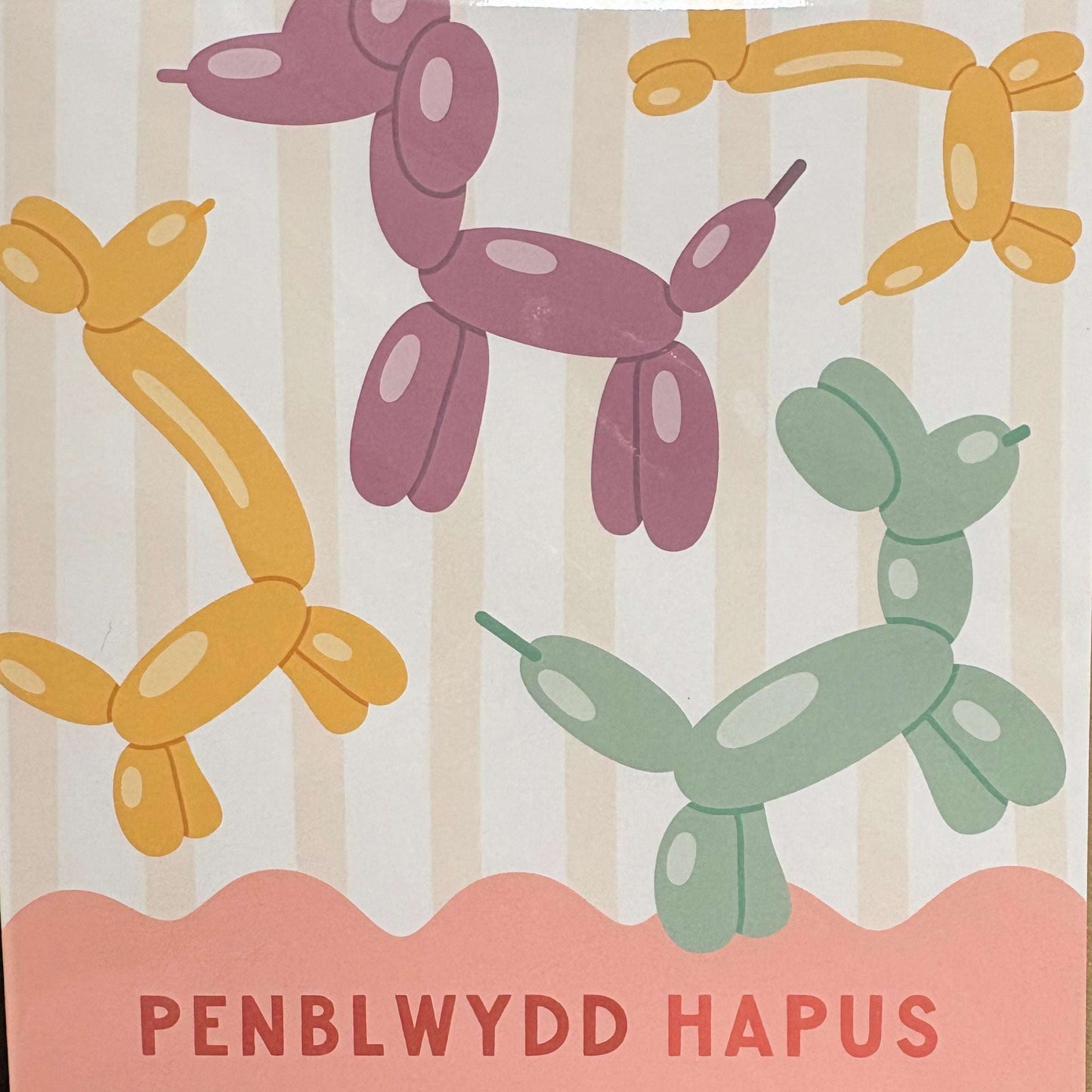 A square greeting card in clear plastic packaging, featuring balloon dog illustrations in yellow, purple, and green on a cream striped background with a peach band reading 'PENBLWYDD HAPUS' in Welsh.