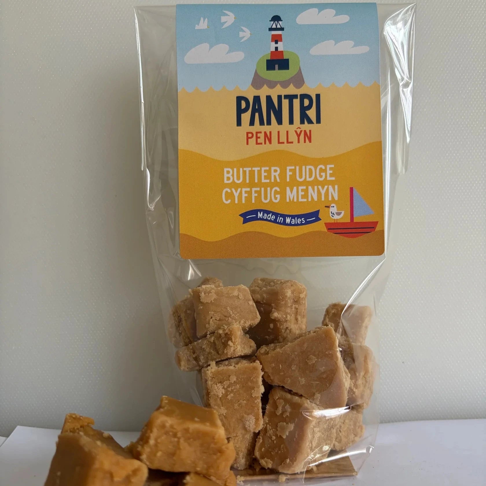 A clear plastic bag of Pantri Pen Llyn Butter Fudge Cffug Menyn with several pieces of golden-brown fudge displayed in front on a white surface against a light gray wall.