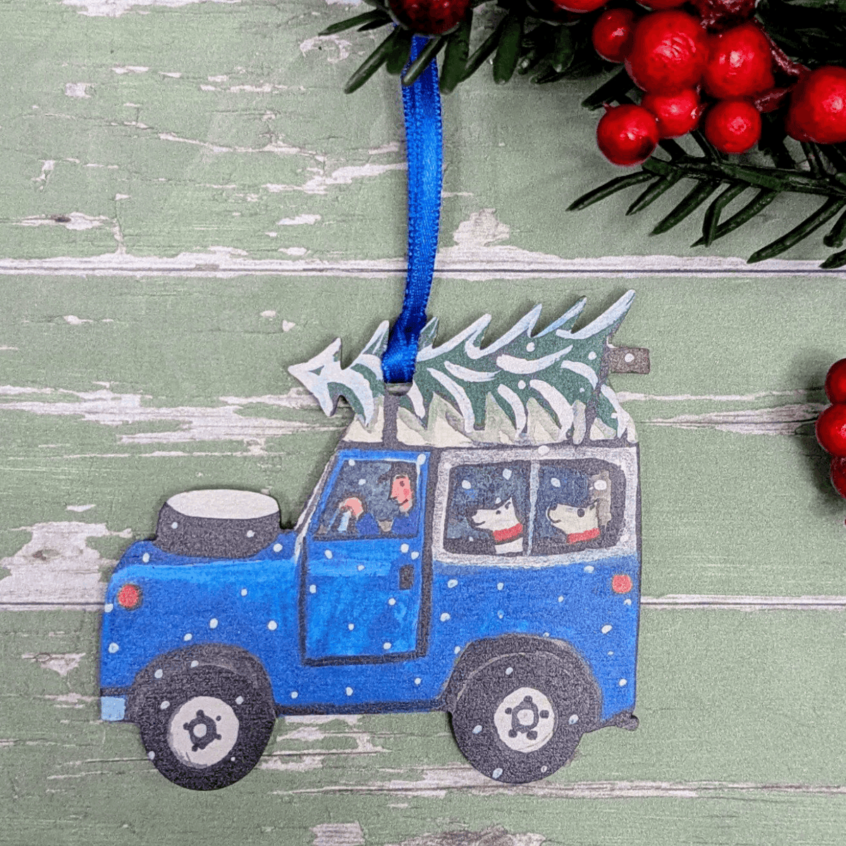 A handcrafted wooden Christmas ornament shaped like a blue car with a tree on top, hanging by a blue ribbon against a distressed green wooden background with red berries and greenery.