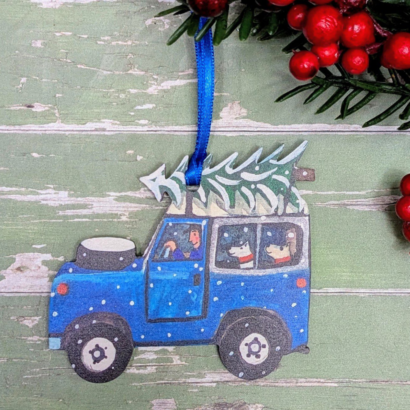 A handcrafted wooden Christmas ornament shaped like a blue car with a tree on top, hanging by a blue ribbon against a distressed green wooden background with red berries and greenery.