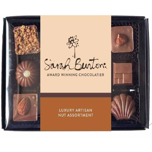 A boxed assortment of chocolates from Sarah Burton, featuring a central cream label with the brand name and a warm brown lower section reading 'Luxury Artisan Nut Assortment.' The black tray contains multiple chocolate shapes, including nuts, caramel, and molded pieces, visible through a clear front window.