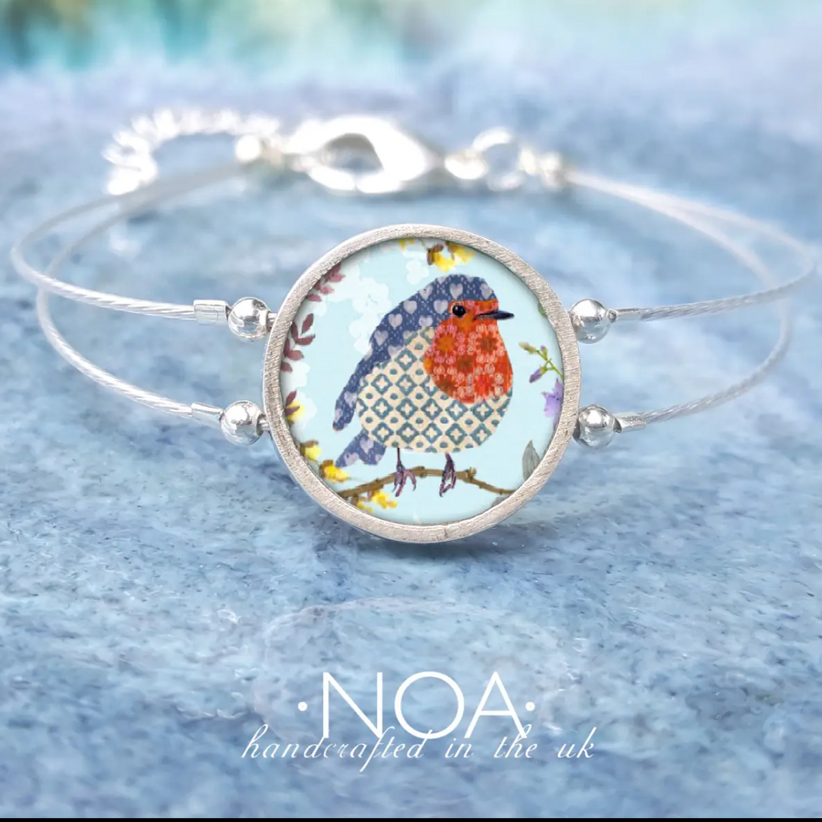 A silver-toned double-band cuff bracelet featuring a round bezel-set cabochon with a colorful robin bird illustration on a light blue background. The brand name 'NOA' is printed below the bracelet.